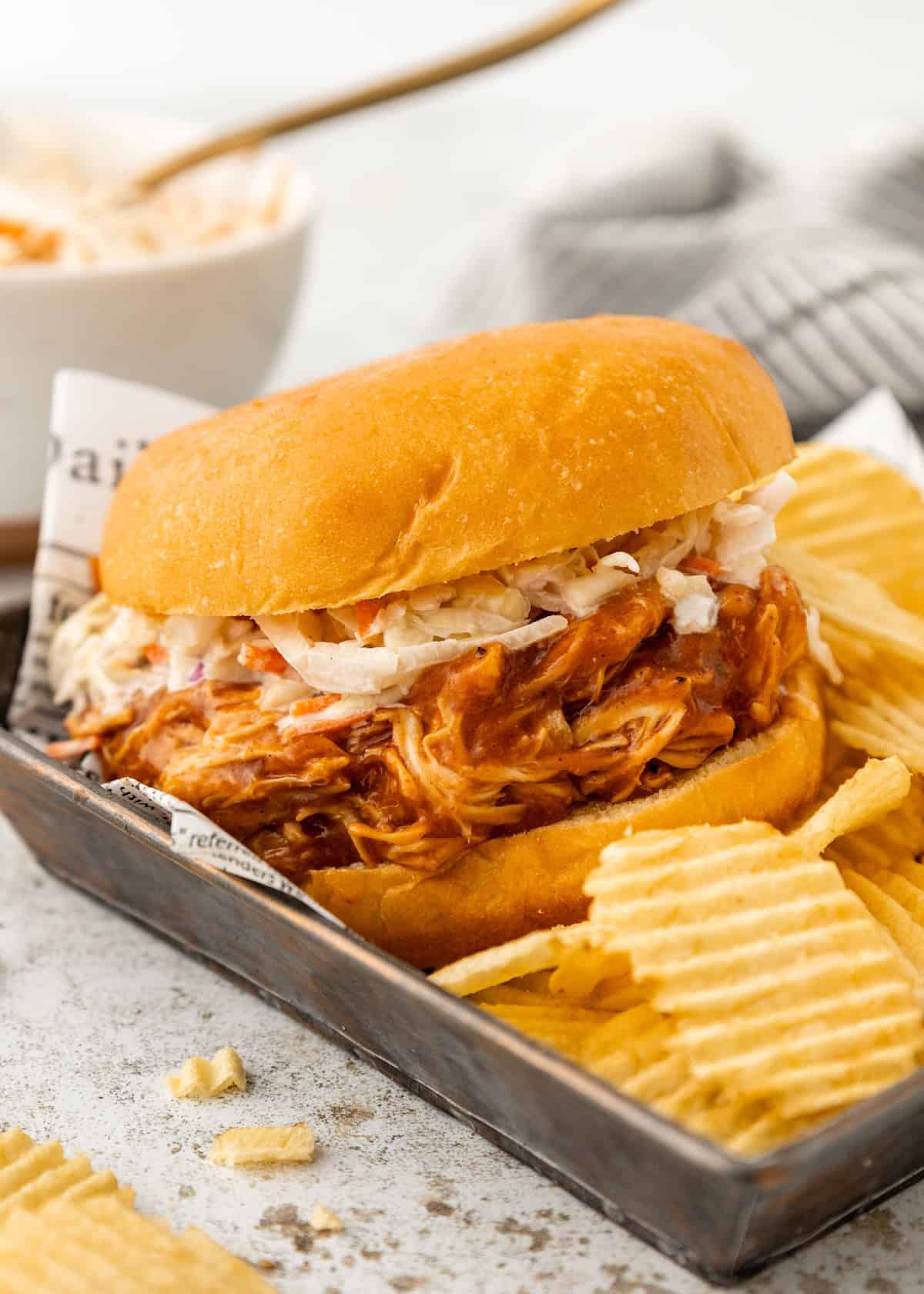 Pulled chicken sandwich with coleslaw on a bun, served with ridged potato chips.