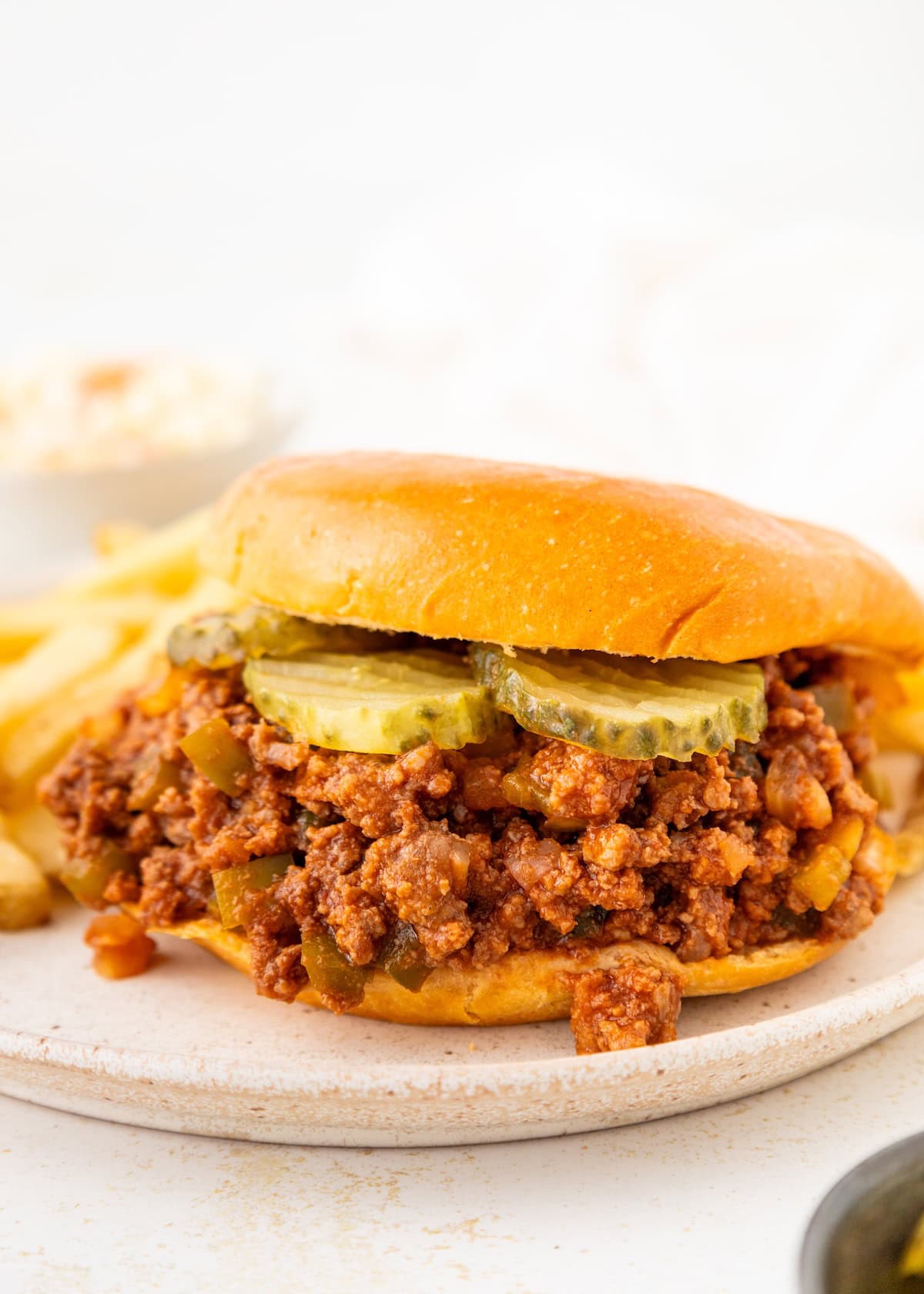 A sloppy joe sandwich with pickles on a plate next to fries and coleslaw.