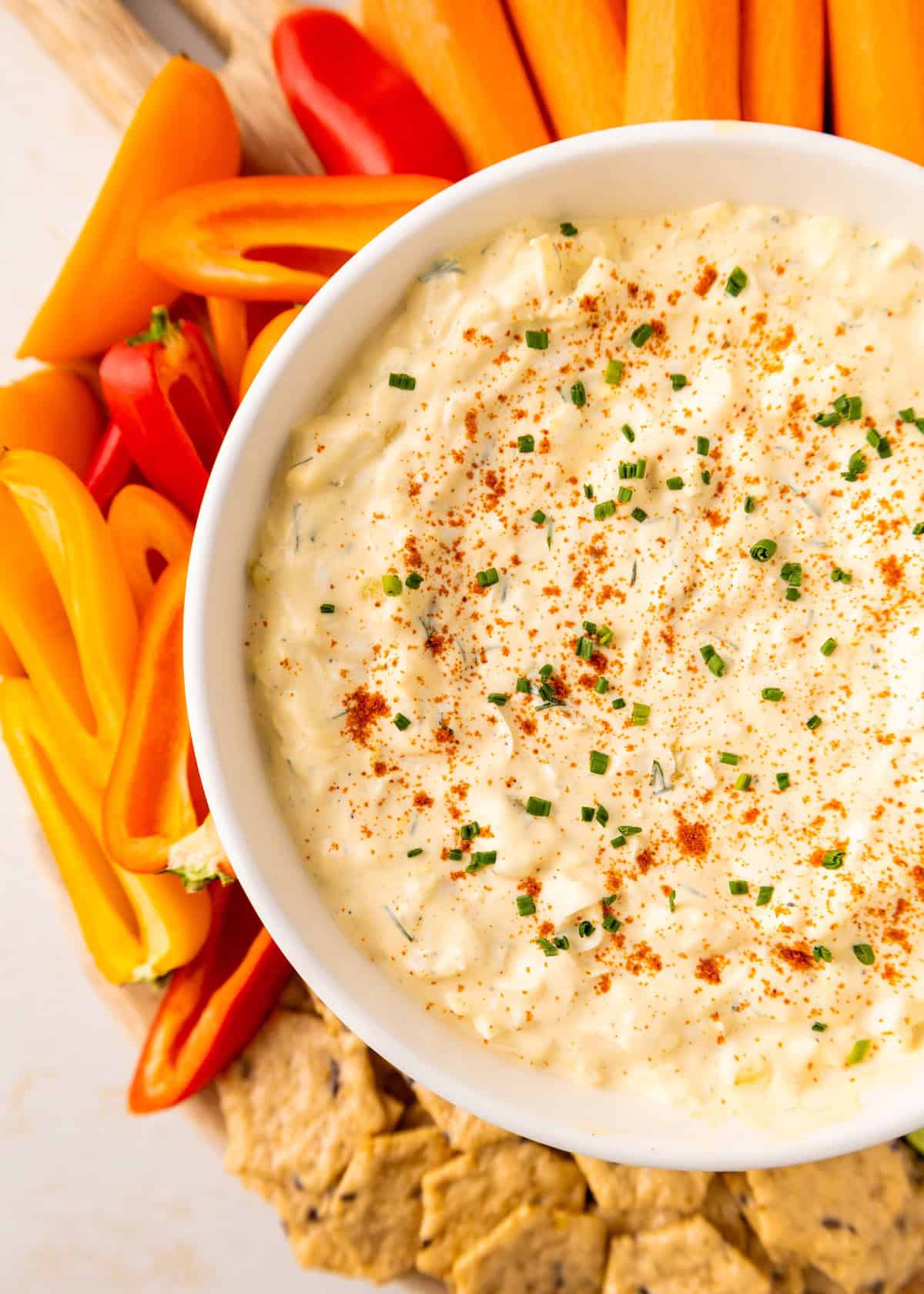 A bowl of creamy dip with chives and paprika, surrounded by peppers, carrots, and crackers.