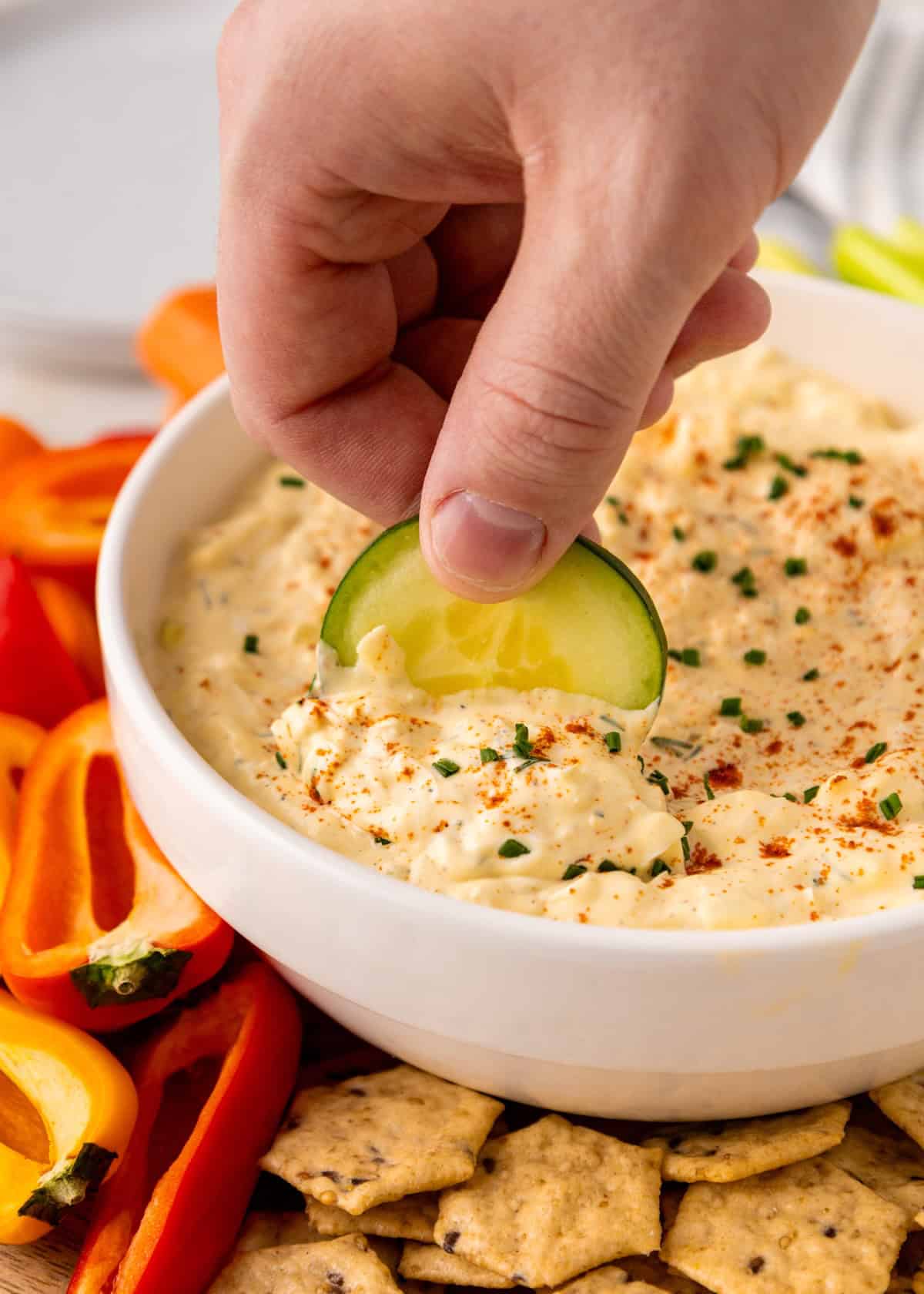 Hand dipping a cucumber slice into a bowl of creamy dip, surrounded by crackers and veggies.