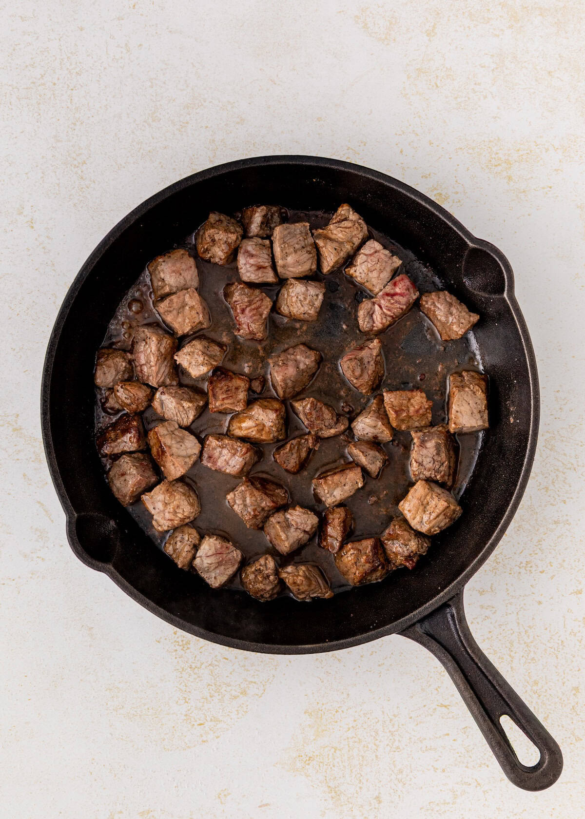 Cubed beef pieces cooking in a black cast iron skillet on a light-colored surface.