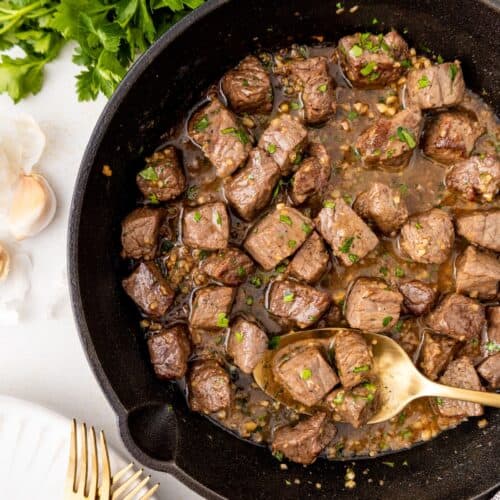 Cubes of cooked steak in a skillet with herbs, next to parsley and garlic on a white surface.
