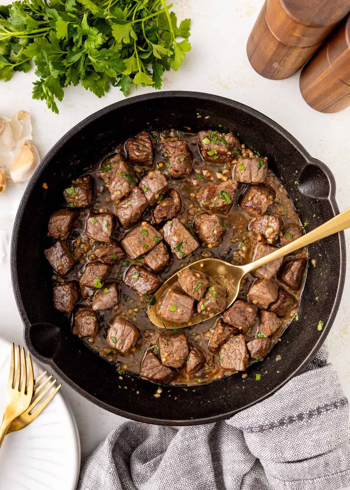 A skillet of garlic butter steak bites garnished with parsley, with a spoon and fresh herbs nearby.