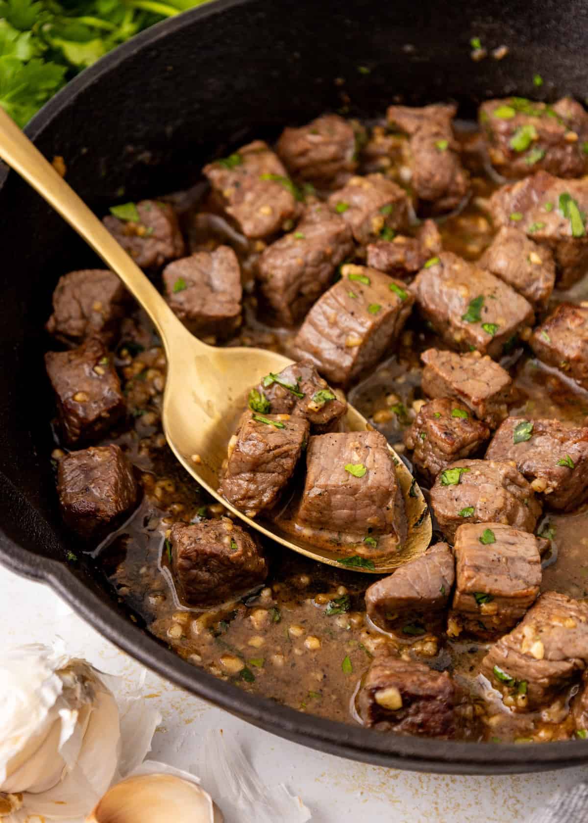 Golden spoon scooping garlic butter steak bites from a cast iron skillet.