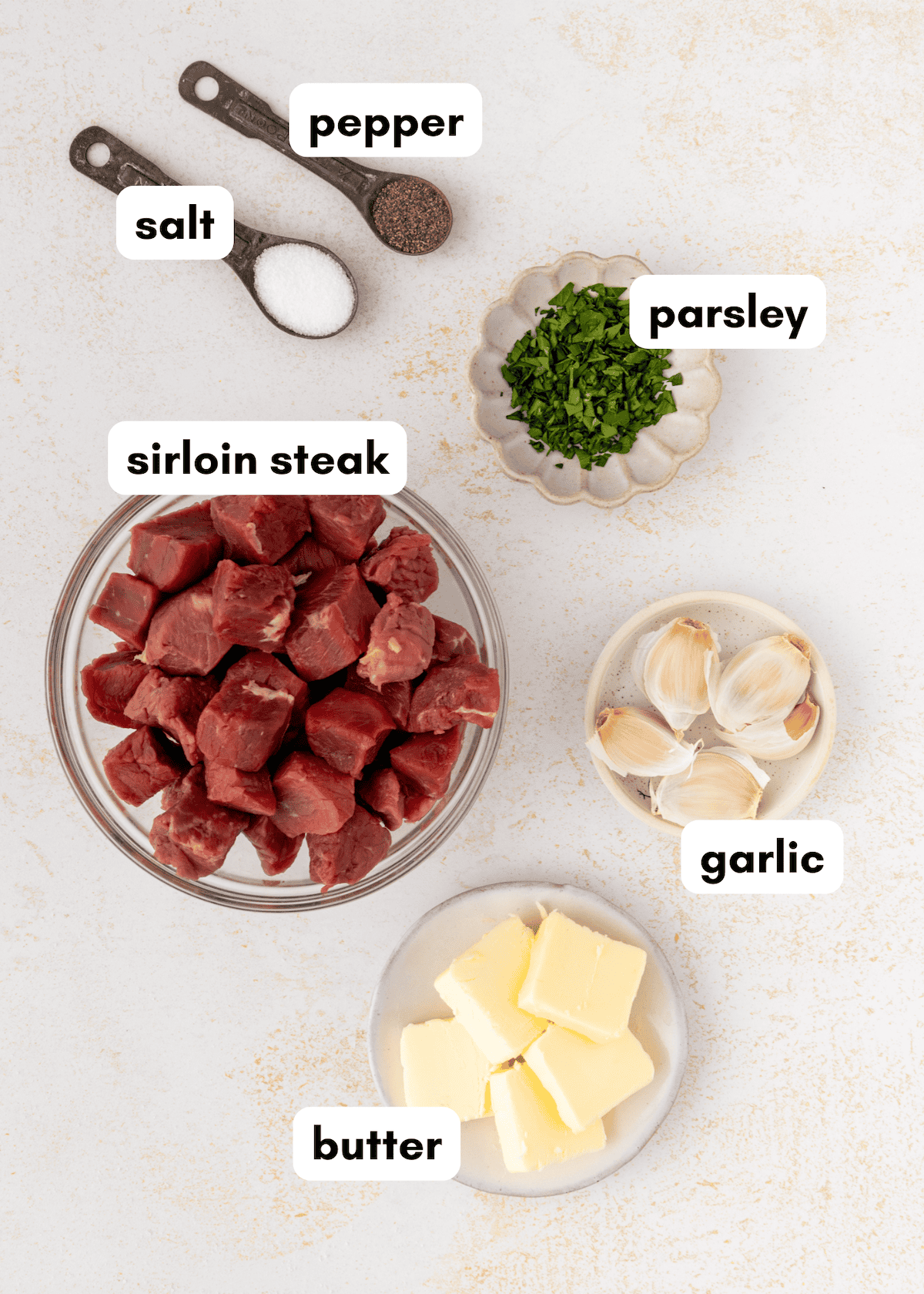 Bowls of garlic butter steak bites with salt, pepper, parsley, and garlic on a light surface.