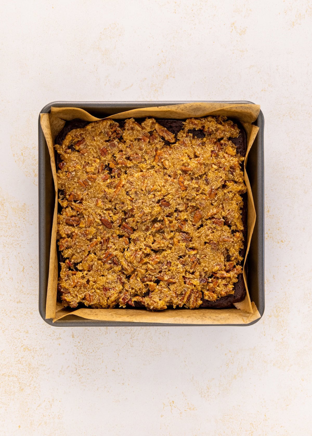 A square baking pan with German chocolate brownies topped with pecan crumble on parchment paper.