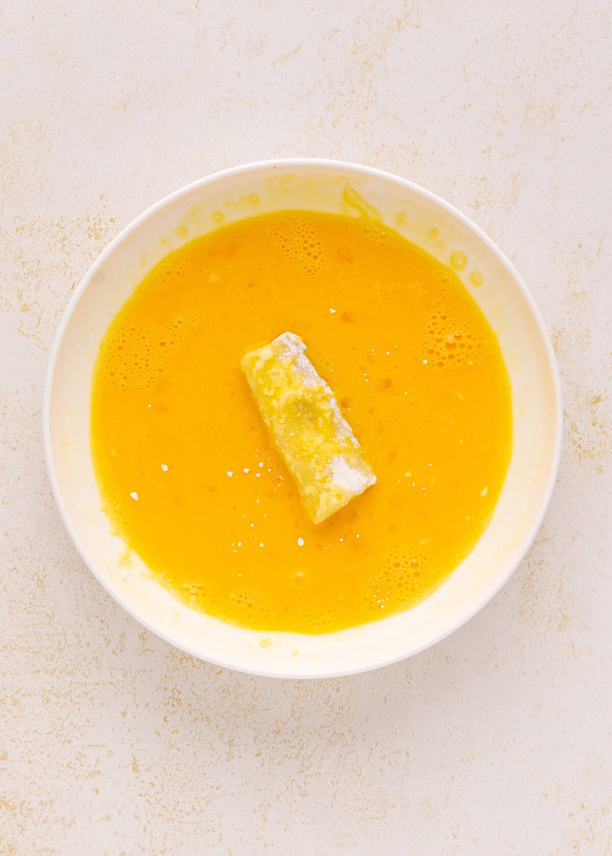 A bowl of beaten eggs with a Homemade Fish Stick partially submerged in the mixture.