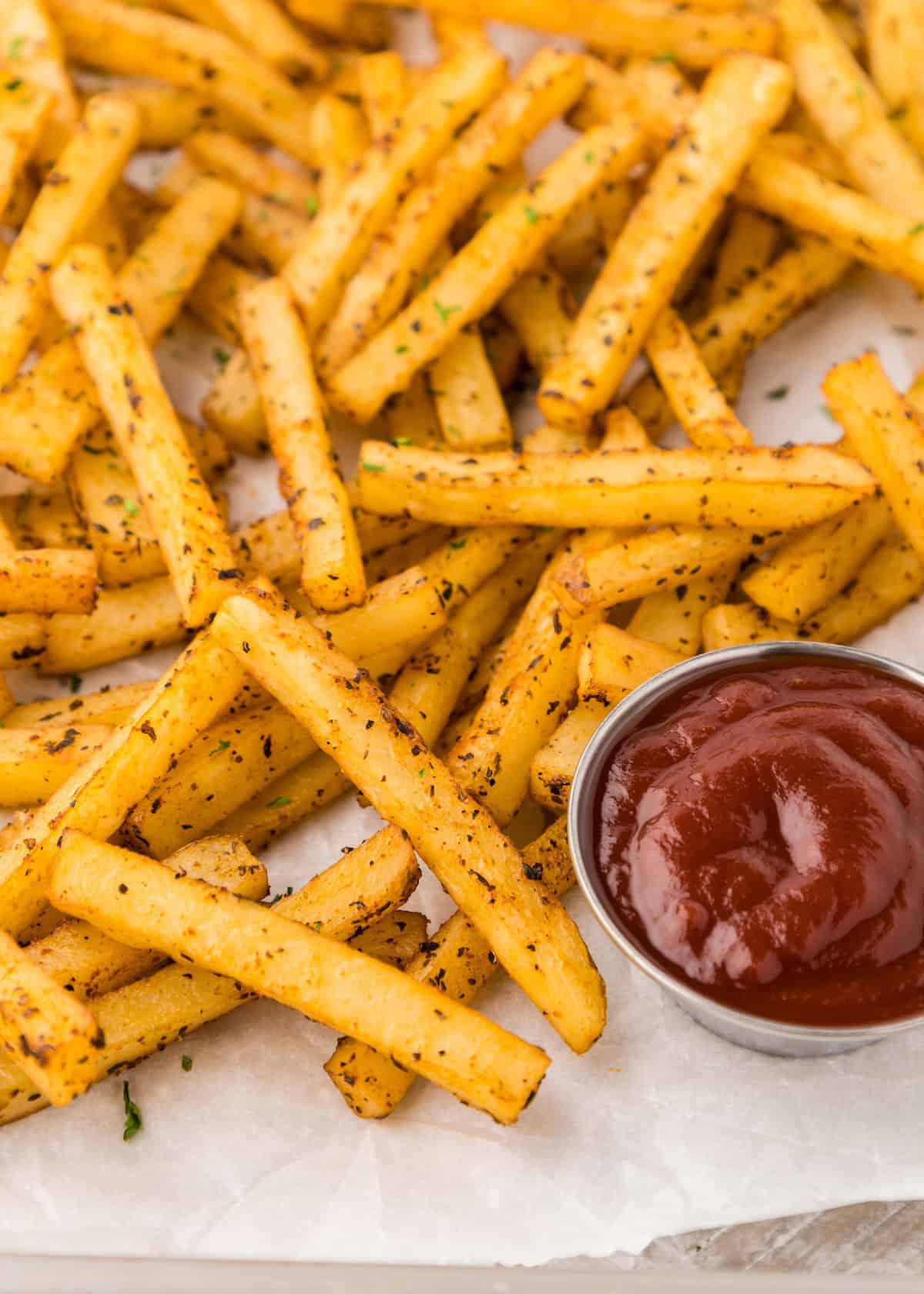 Seasoned French fries with herbs next to a small metal cup of ketchup on parchment paper.