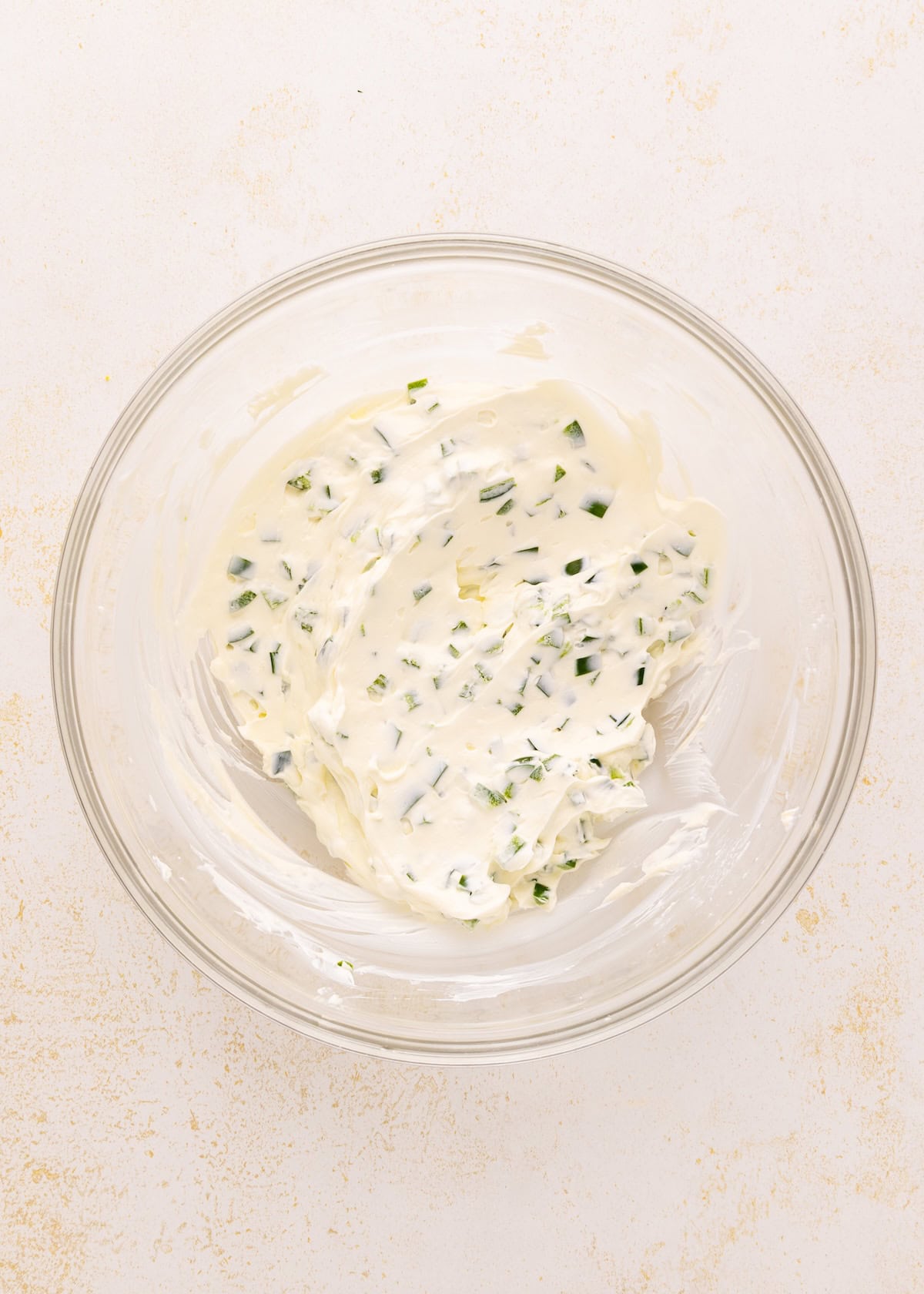 Glass bowl with creamy dip mixed with chopped green herbs on a light surface.