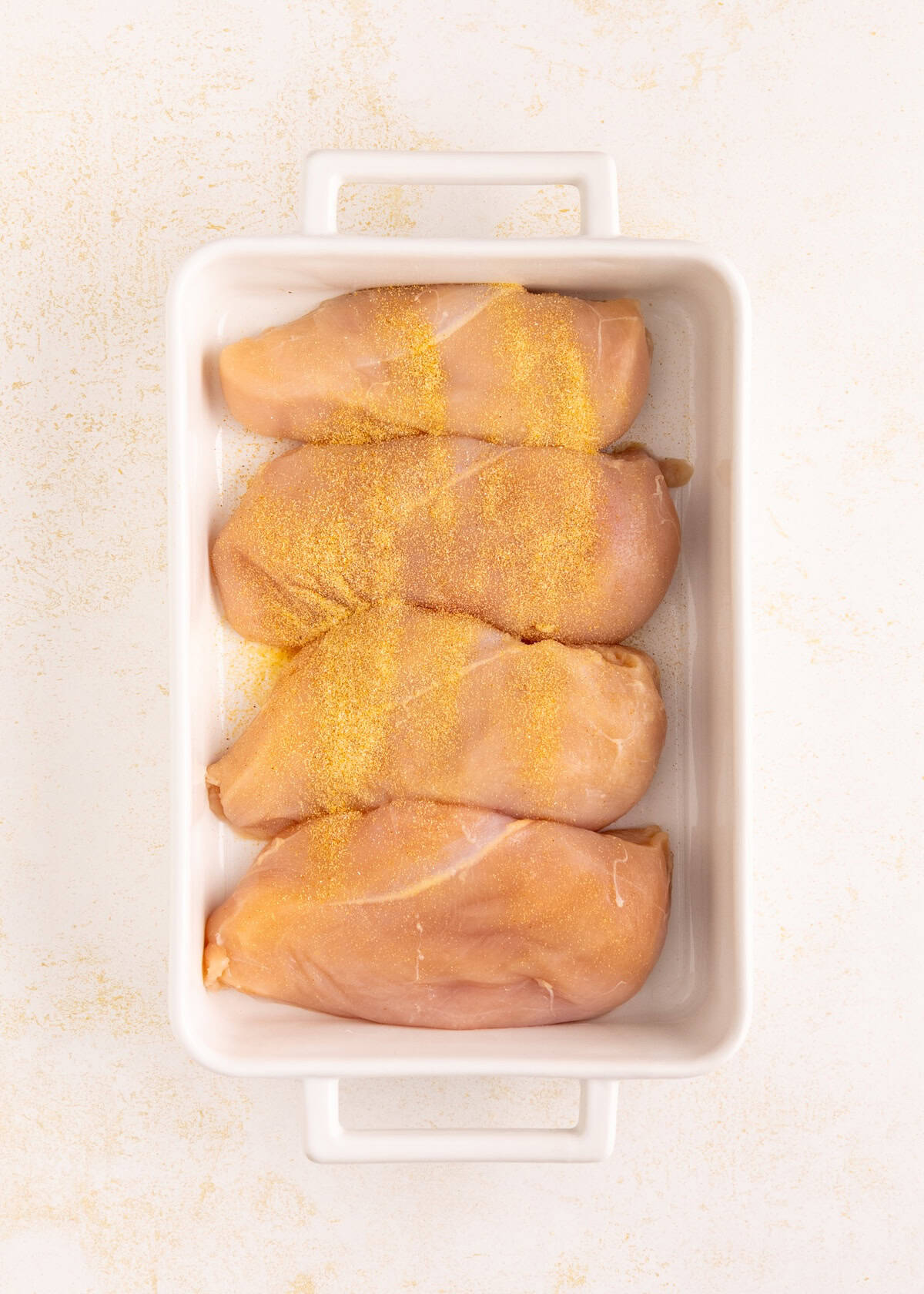 Four raw chicken breasts topped with seasoning in a white rectangular baking dish.