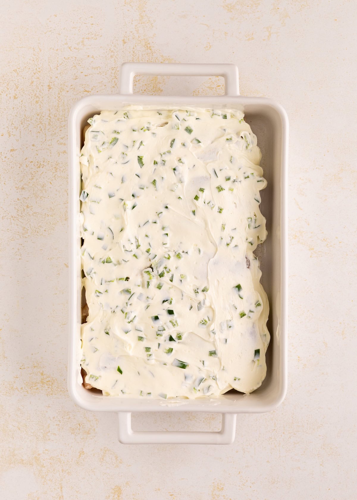 Rectangular dish with a creamy mixture topped with chopped green herbs, ready to be baked.