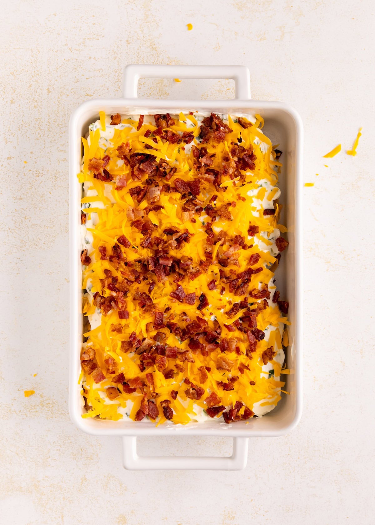 Rectangular dish with shredded cheese and crumbled bacon on top of a creamy white layer.