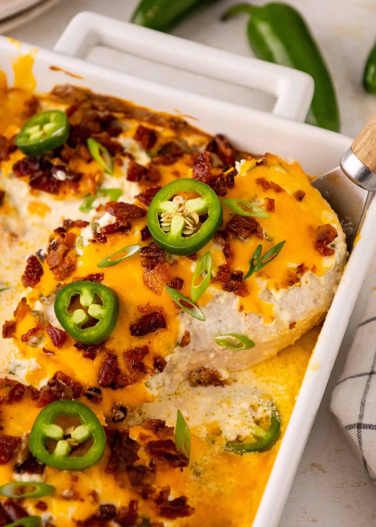 Casserole topped with melted cheese, bacon, jalapeño slices, and green onions in a white dish.