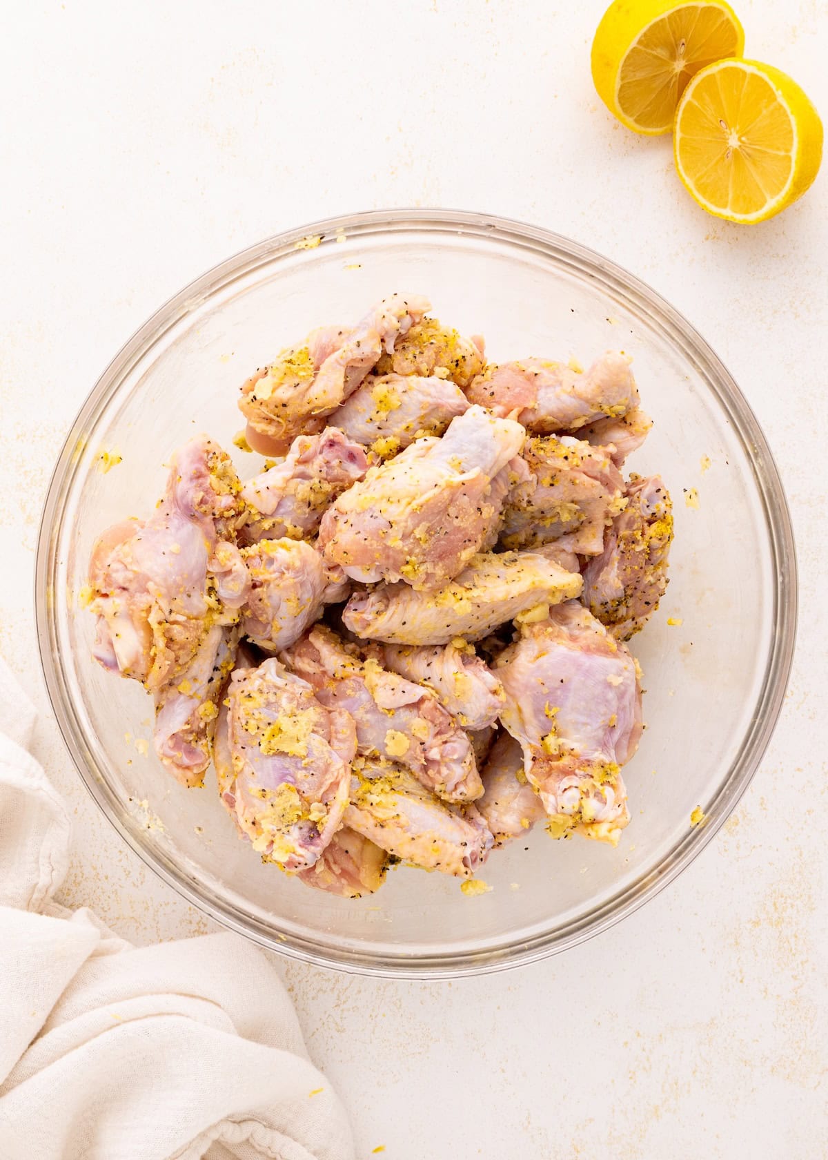 Raw chicken wings marinated with spices in a glass bowl, with lemon halves beside it.