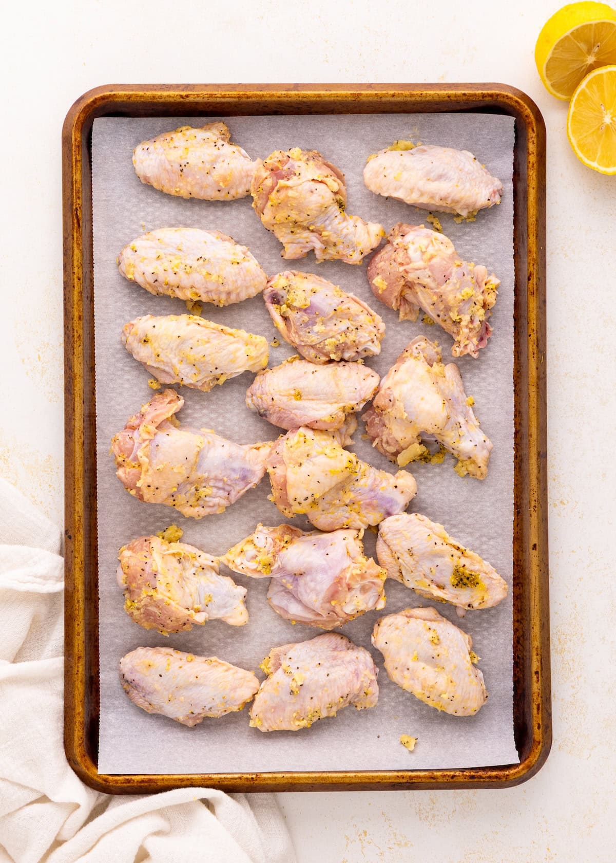 Raw, seasoned chicken wings on a baking sheet with lemon halves in the corner.