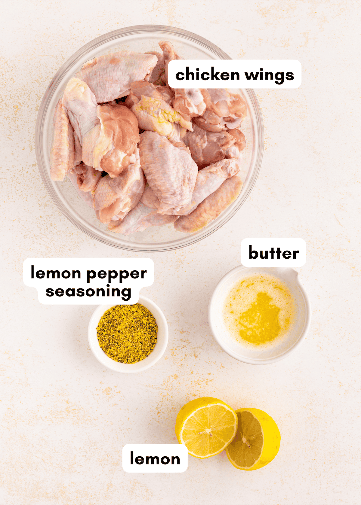 Raw chicken wings, lemon, melted butter, and lemon pepper seasoning in separate bowls on a table.