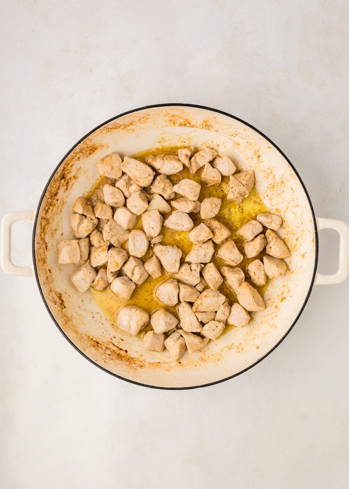 Cubed chicken pieces cooking in a white, round skillet with handles on a light surface.