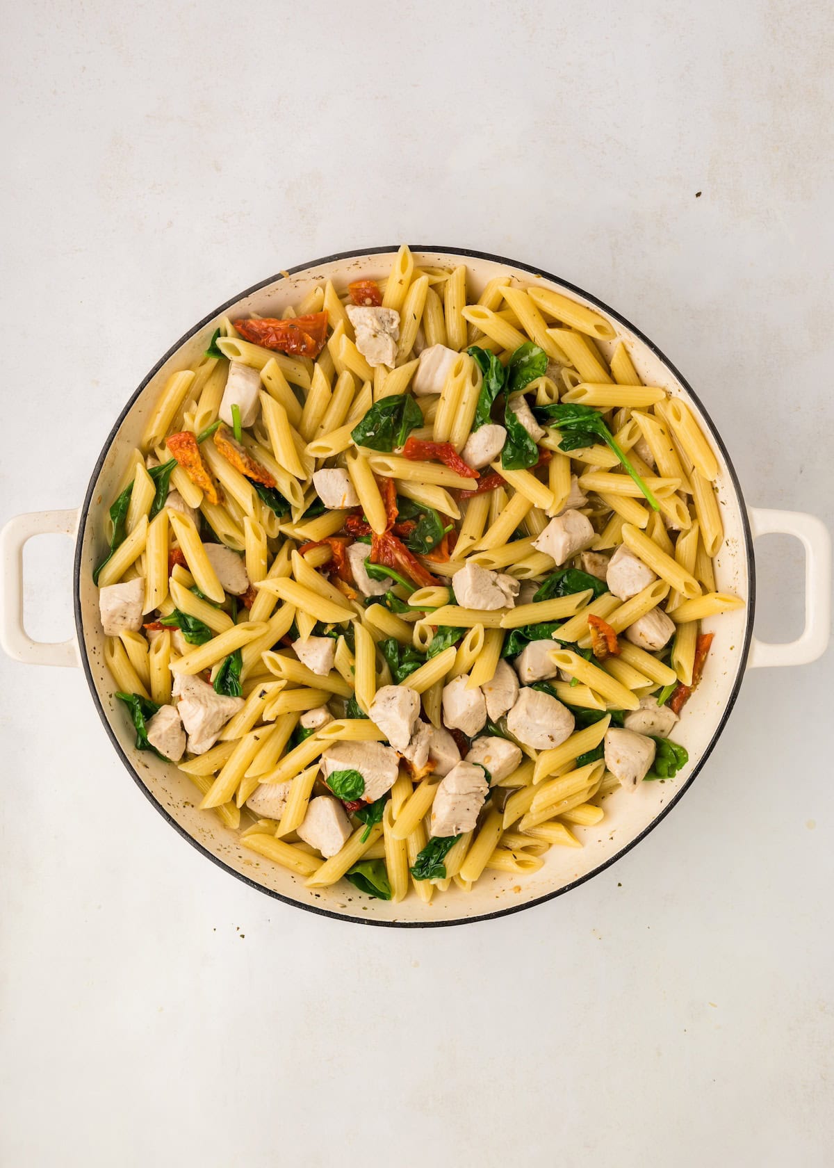 Penne pasta with chicken, spinach, and sun-dried tomatoes in a white handled pan.