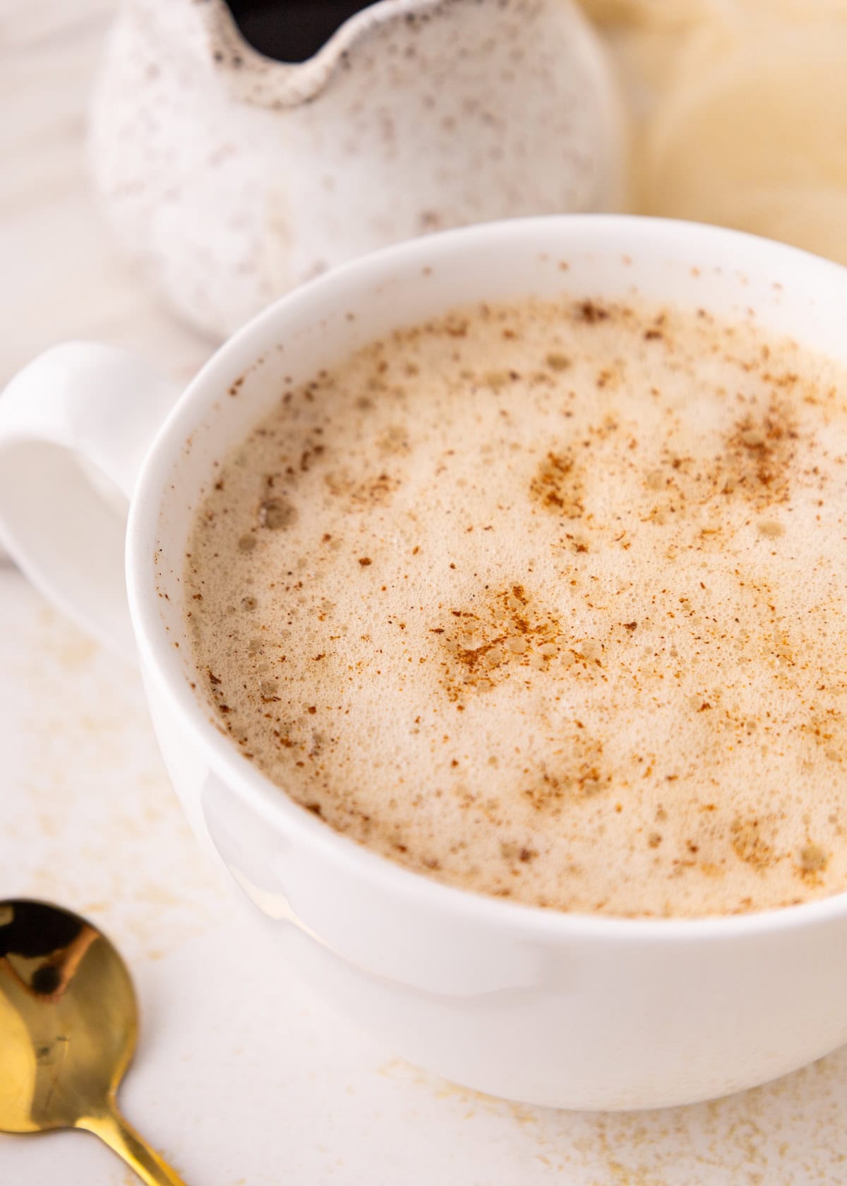 A frothy chai latte in a white cup, sprinkled with cinnamon, next to a gold spoon.
