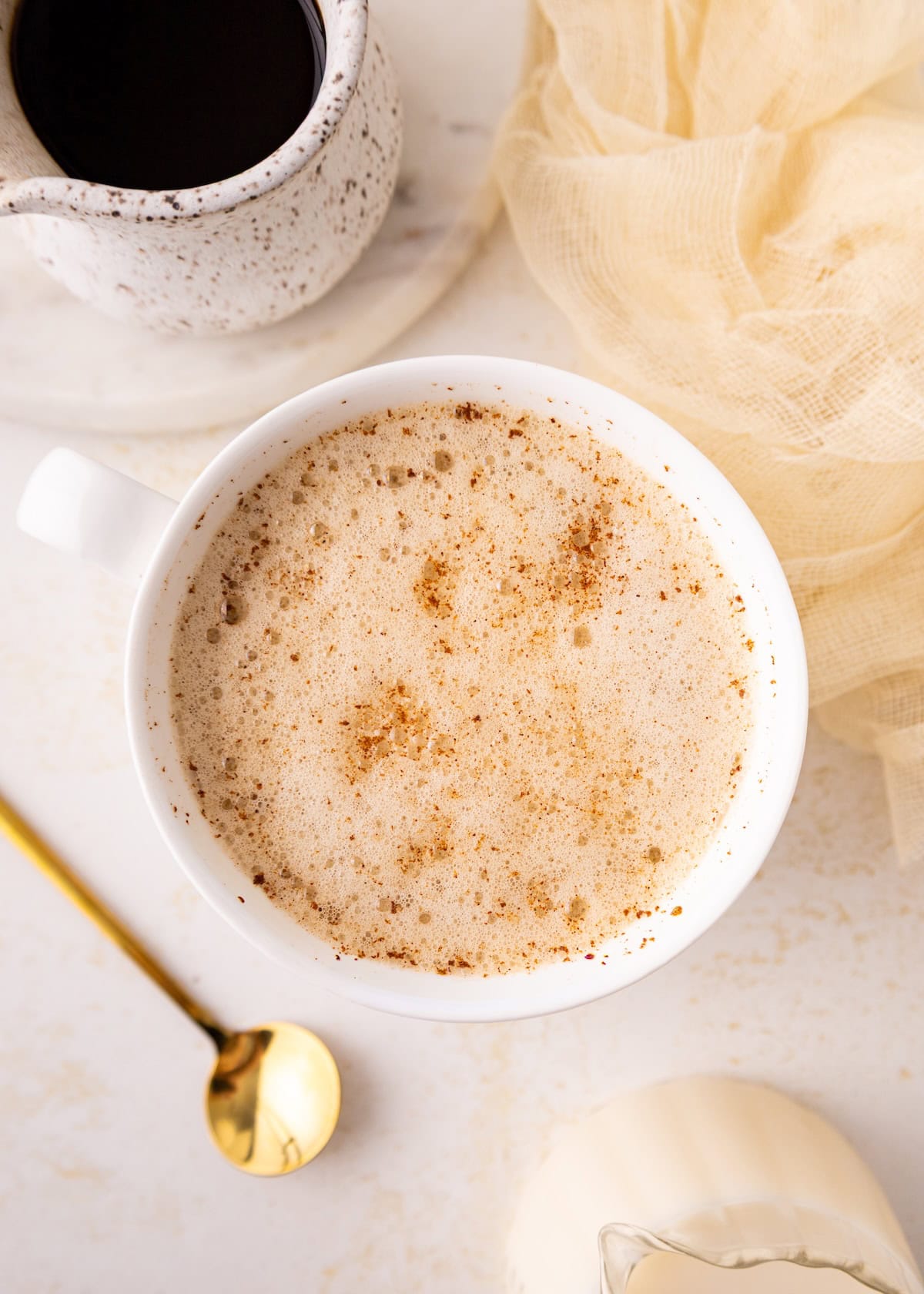 A frothy cup of chai latte with cinnamon, next to a gold spoon and a small cream pitcher.