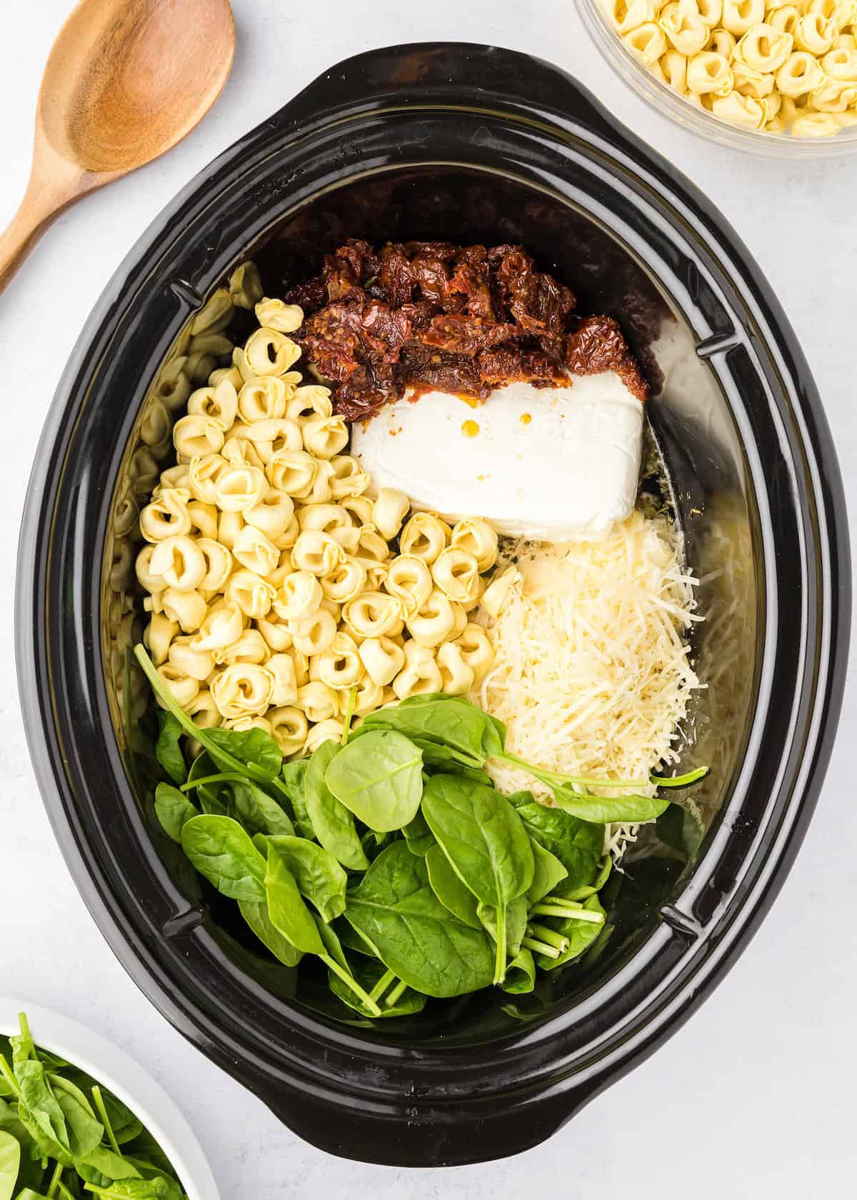 Crockpot with spinach, tortellini, sun-dried tomatoes, ricotta, and shredded cheese inside.