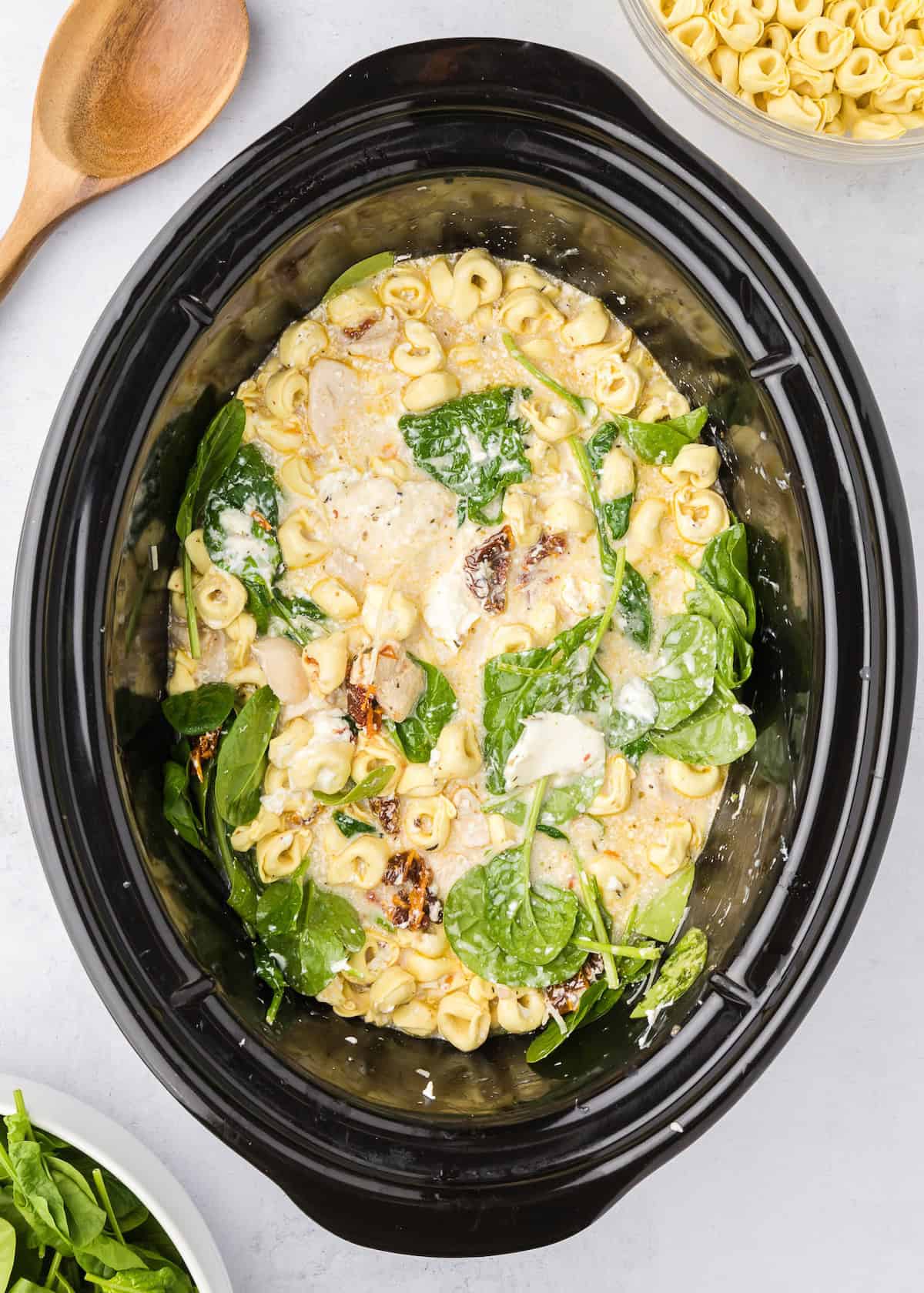 Creamy tortellini, spinach, and chicken in a slow cooker, with a wooden spoon nearby.