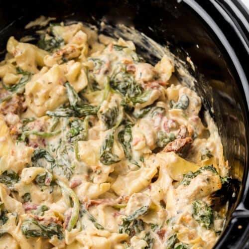 Creamy pasta with spinach and sun-dried tomatoes in a black slow cooker.