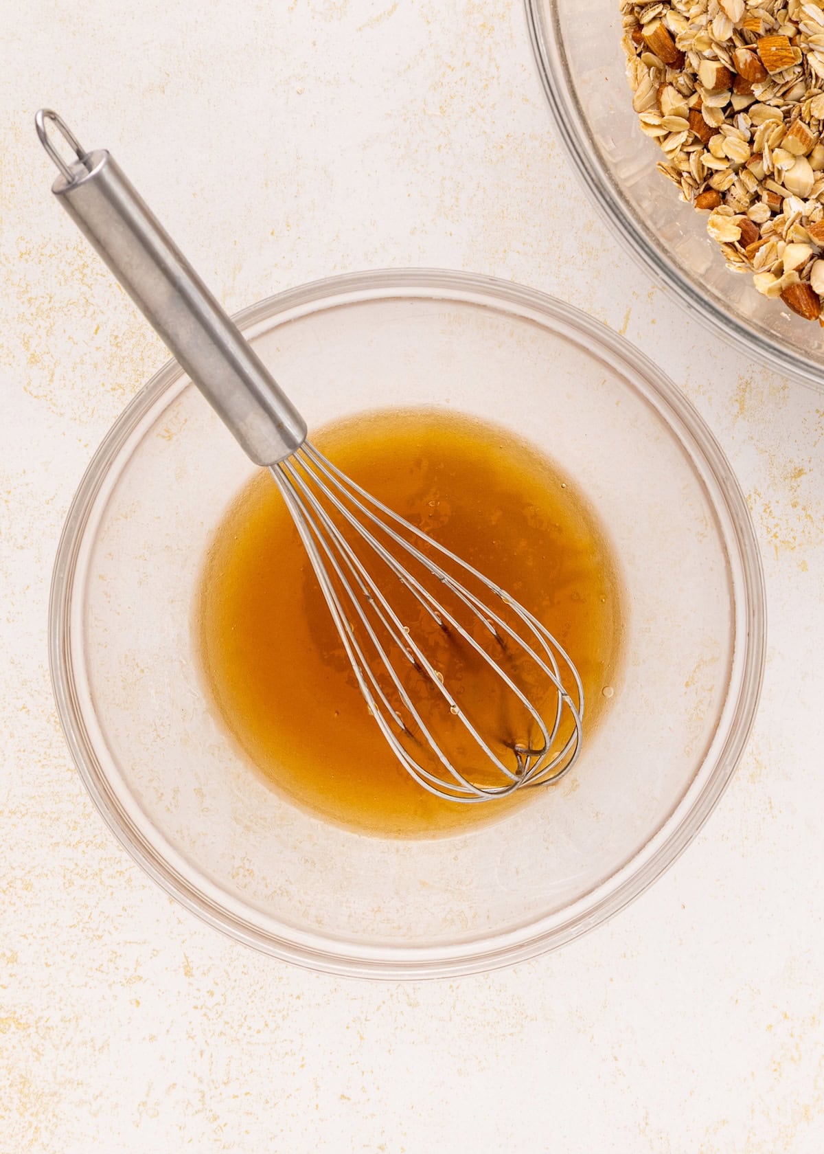 A whisk in a bowl of brown liquid next to oats and nuts for Vanilla Almond Granola.