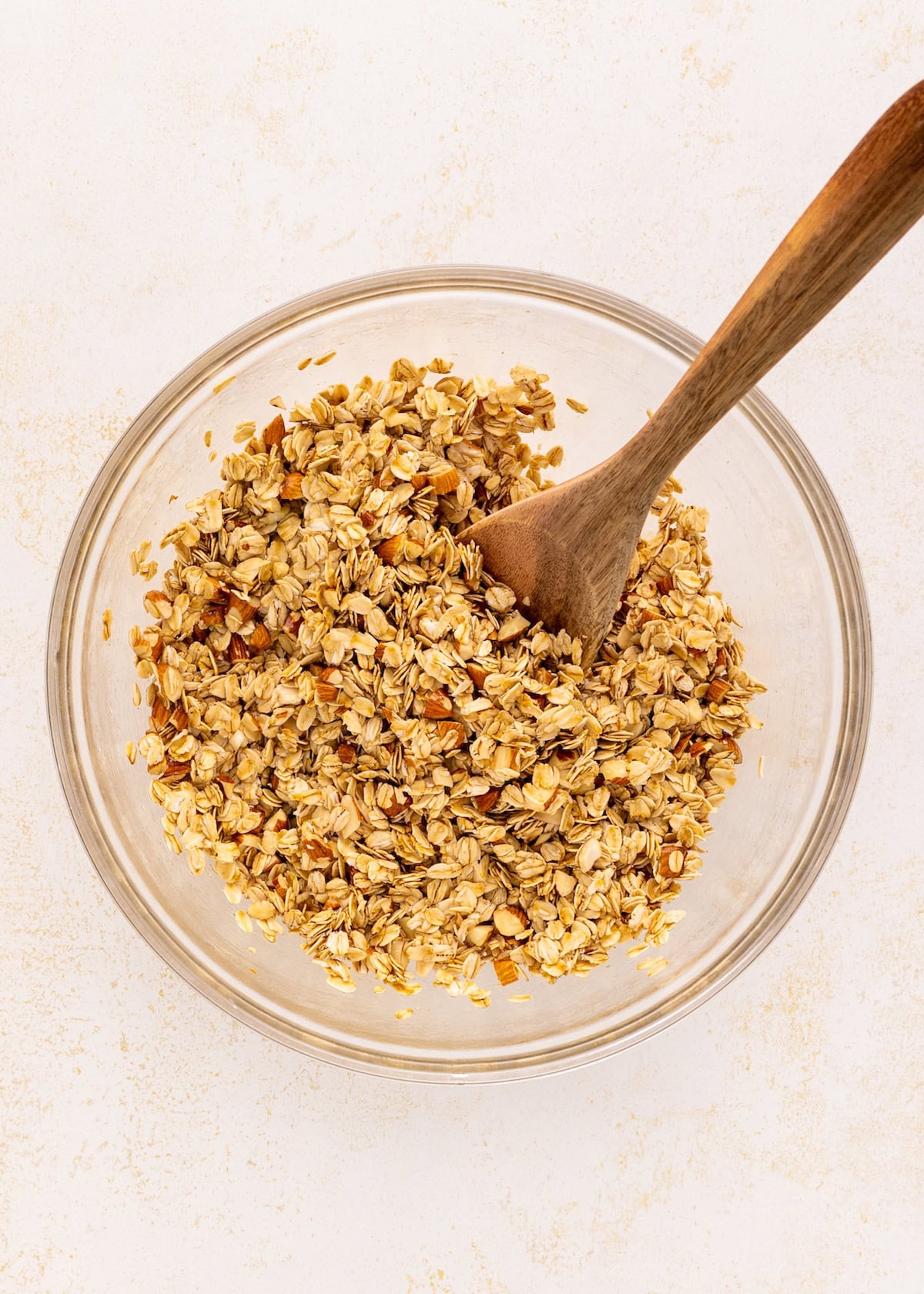 A glass bowl of Vanilla Almond Granola with a wooden spoon on a light surface.