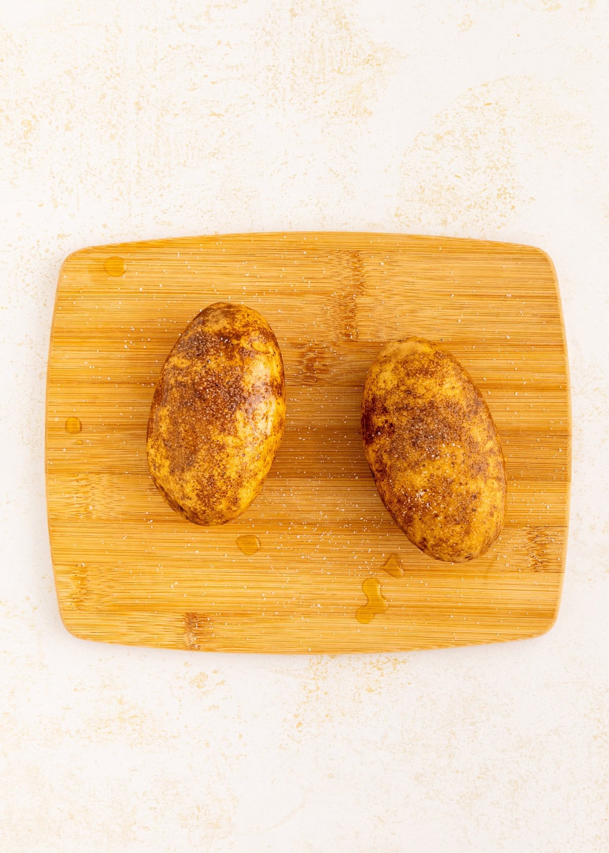 Two seasoned potatoes on a wooden cutting board with a few drops of oil.