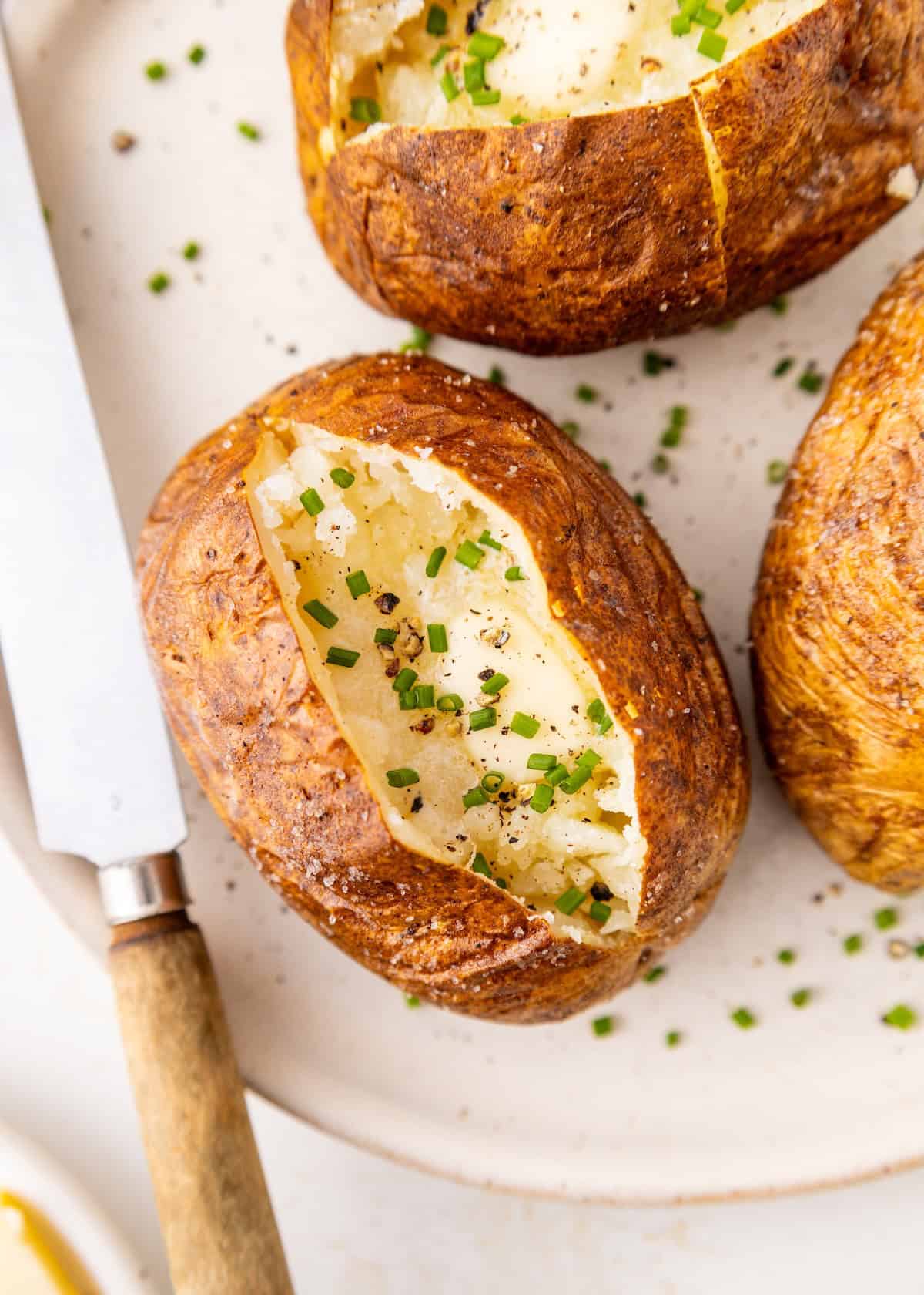 A baked potato with chives and pepper, split open on a plate with a knife beside it.