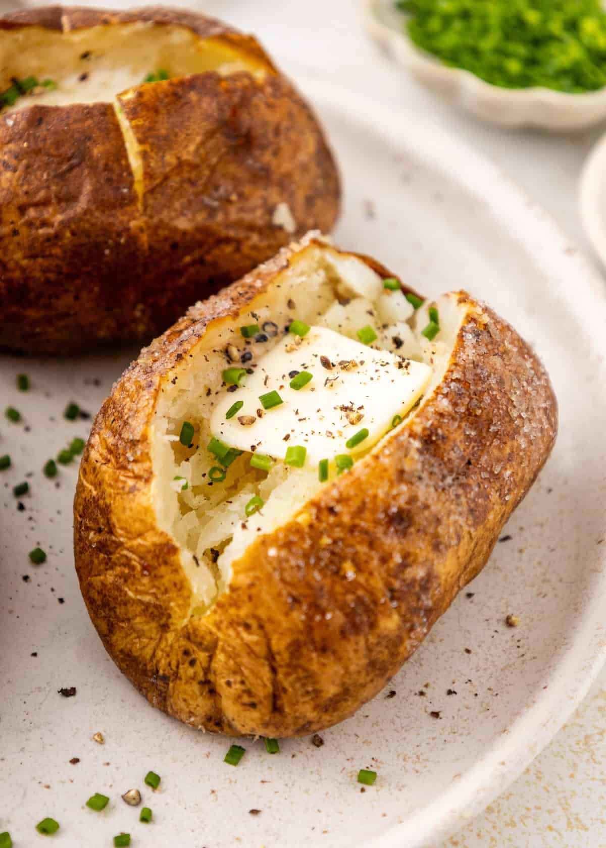 A baked potato with butter, chives, and pepper on a white plate.