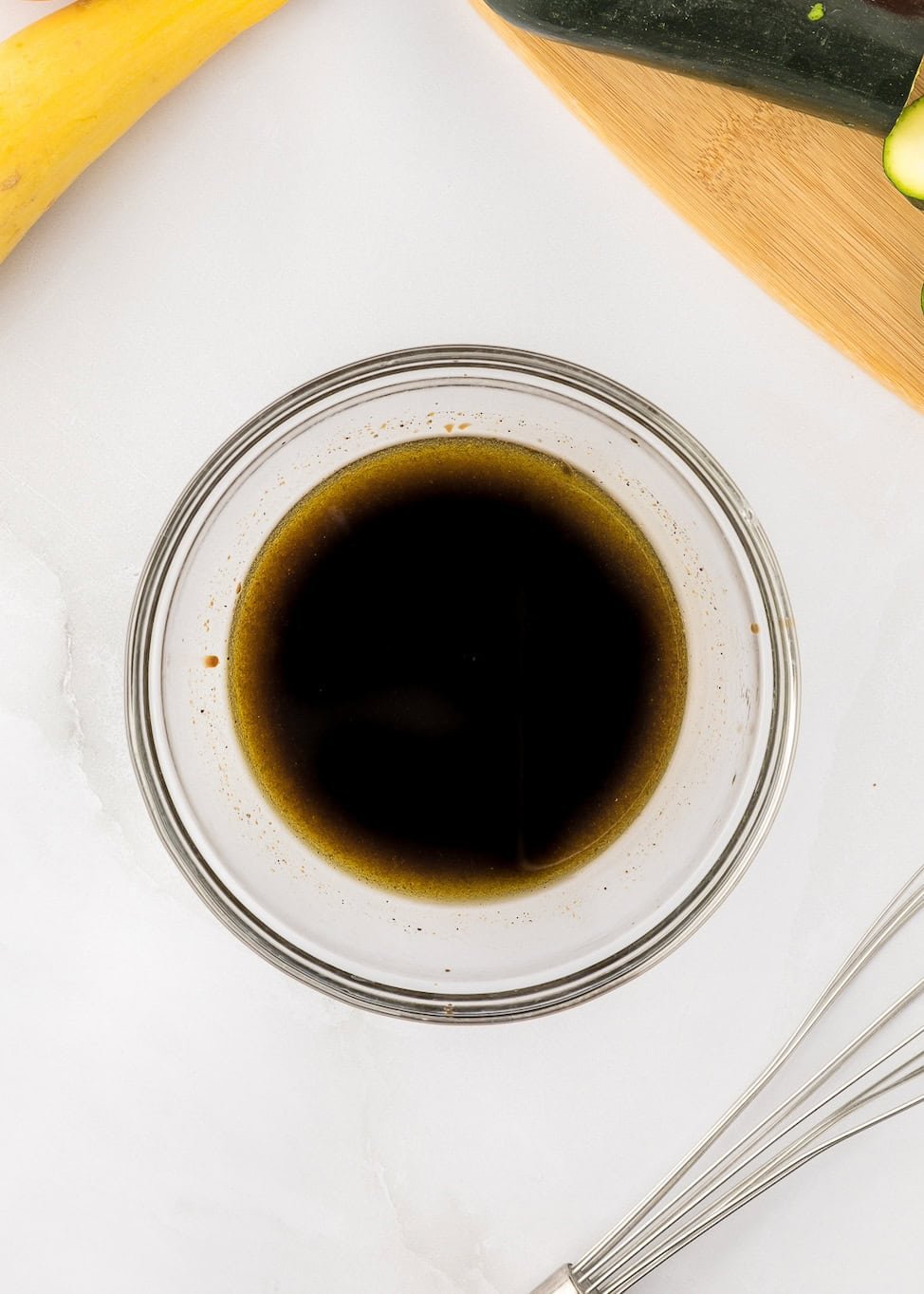 A glass bowl of dark liquid on a white surface with a whisk and vegetables nearby.