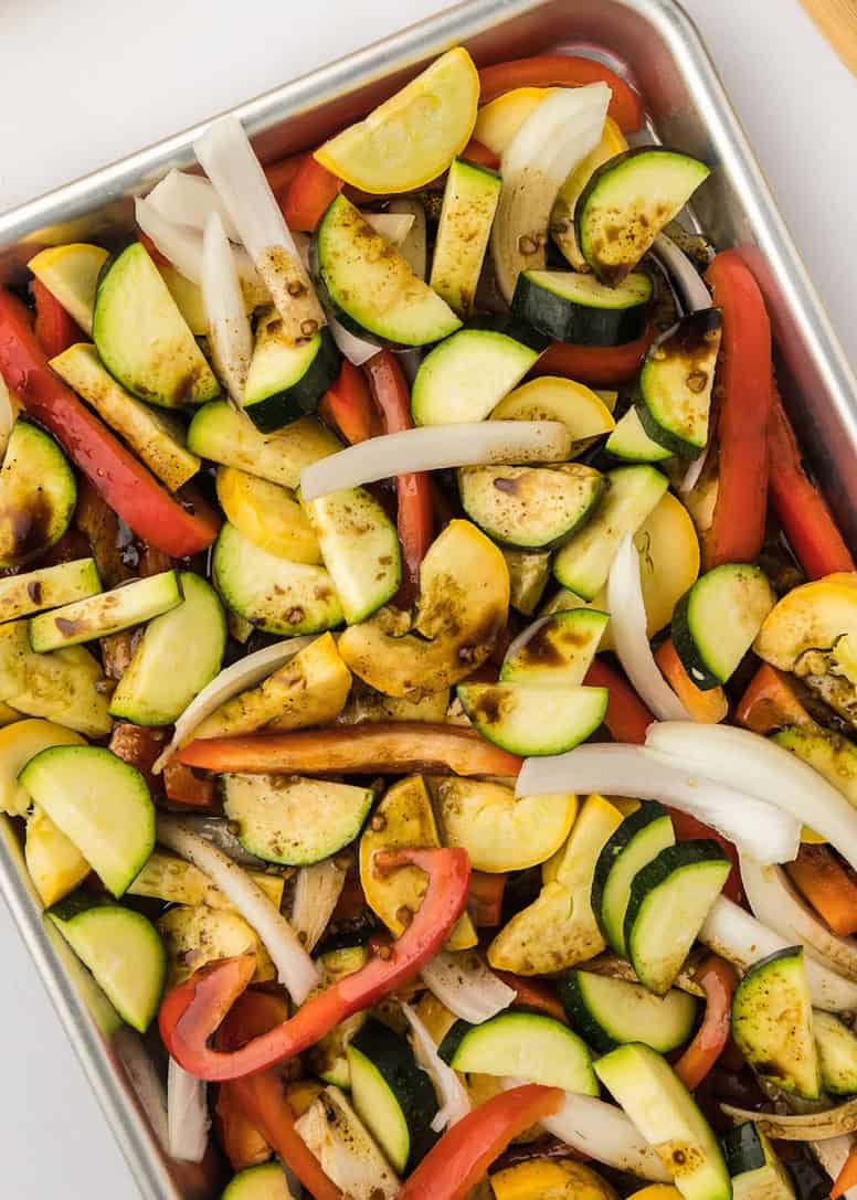 Sliced zucchini, squash, onion, and red bell pepper on a baking sheet, seasoned for roasting.