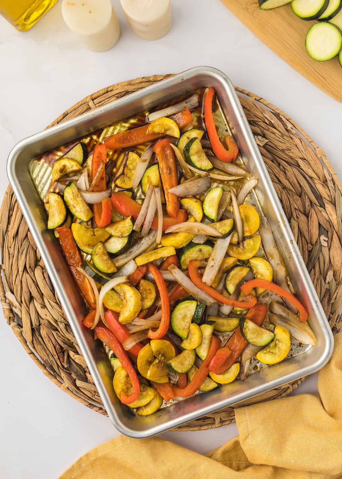 Roasted mixed vegetables in a metal tray on a woven placemat, with zucchini and peppers.