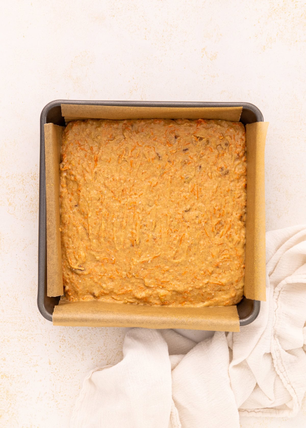 Square baking pan with carrot cake batter, lined with parchment, on a light surface.