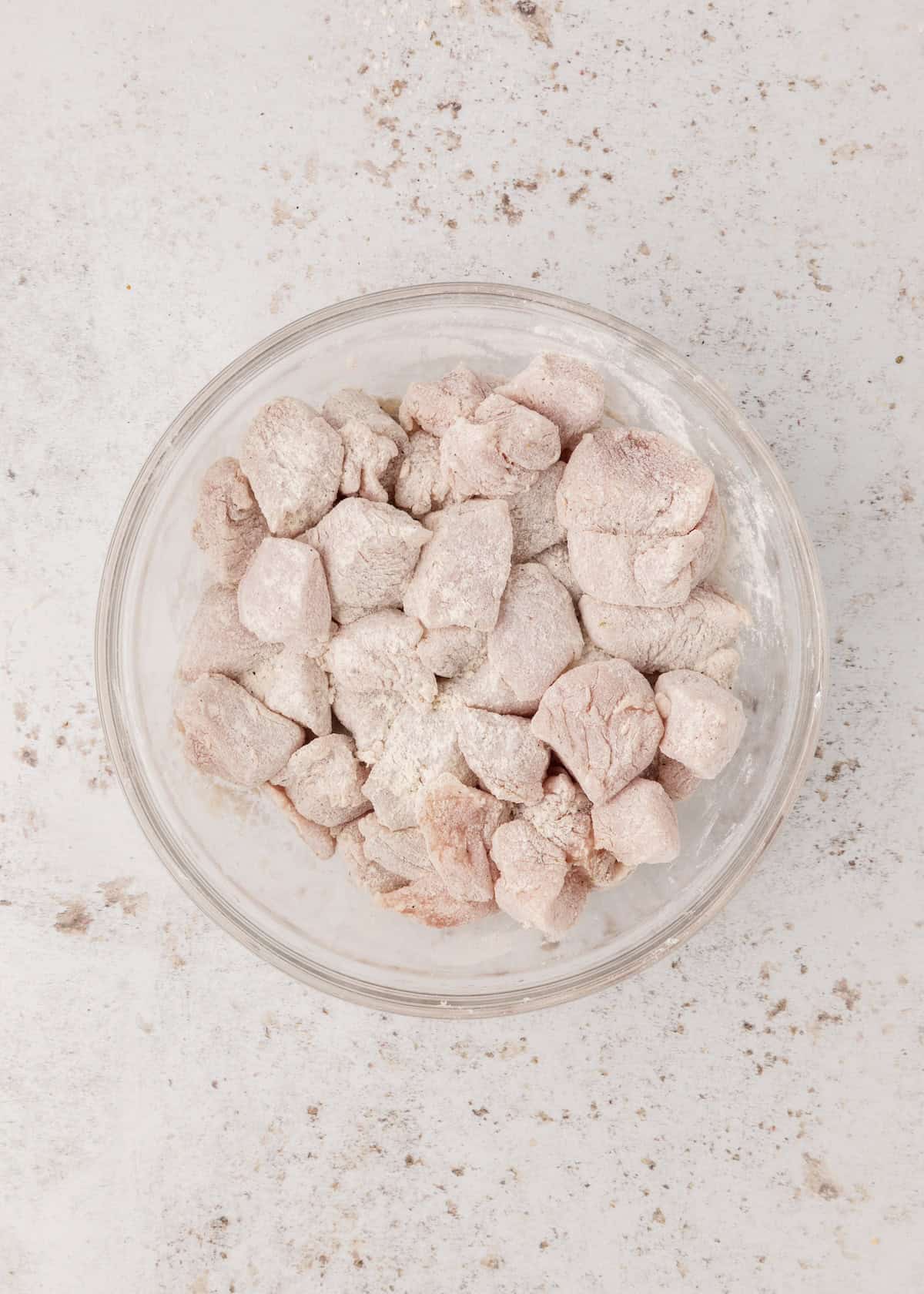 A glass bowl filled with flour-coated raw meat chunks on a light surface.