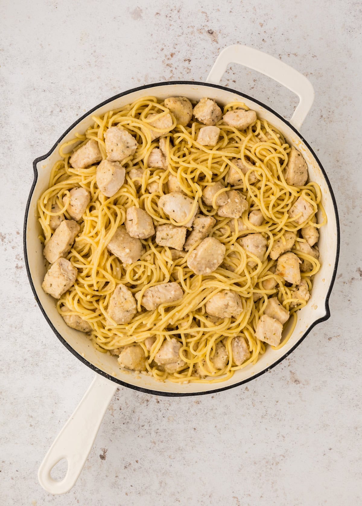 A white skillet filled with spaghetti and chunks of cooked chicken on a light background.