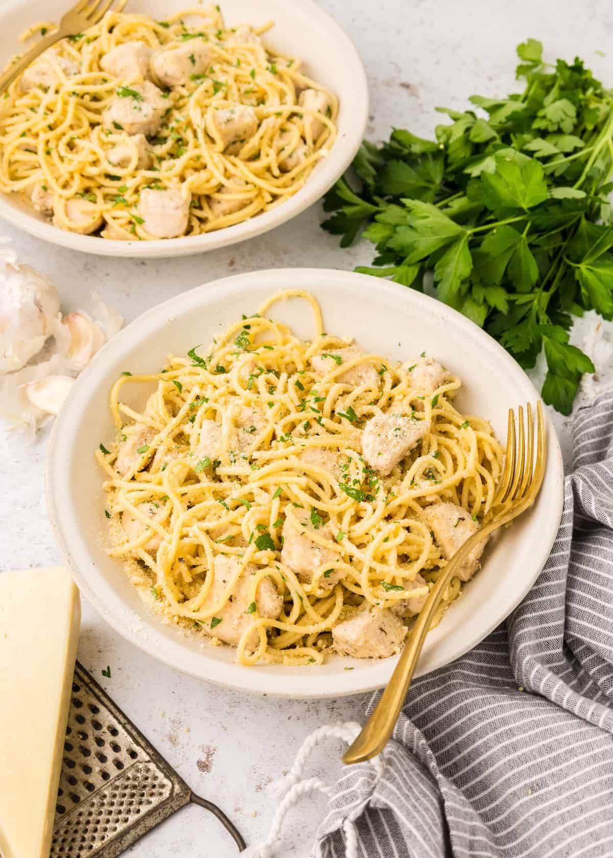 Two bowls of creamy chicken pasta with parsley, cheese, and a gold fork on a white table.
