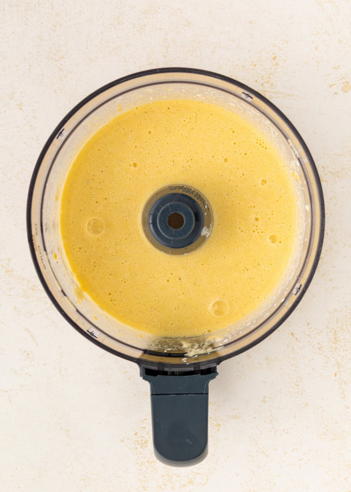 Top view of a food processor with yellow batter inside on a light surface.