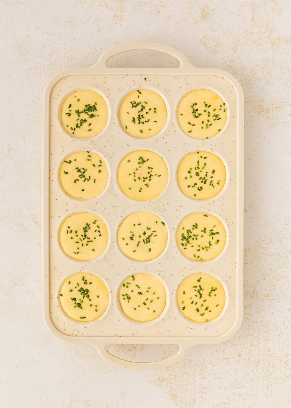 A baking tray with twelve round egg bites topped with chopped green herbs.