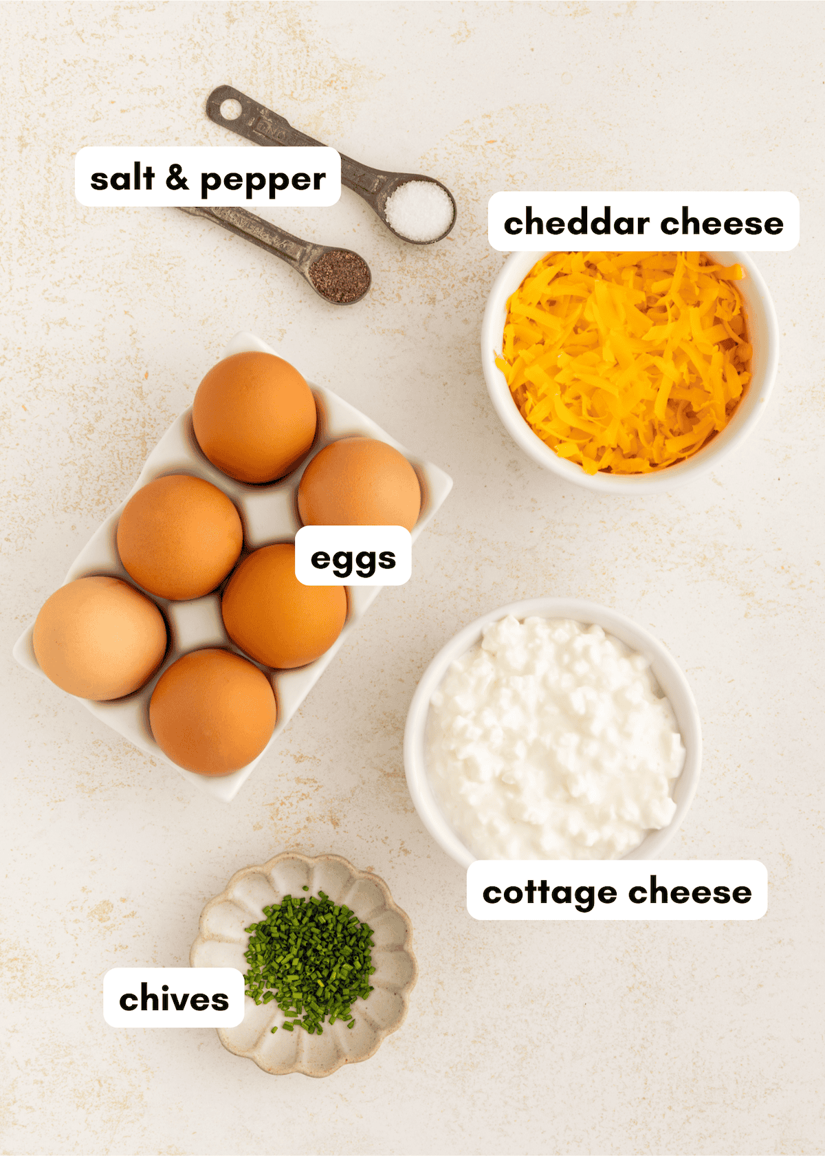 Eggs, chives, salt & pepper, shredded cheddar cheese, and cottage cheese on a light surface.
