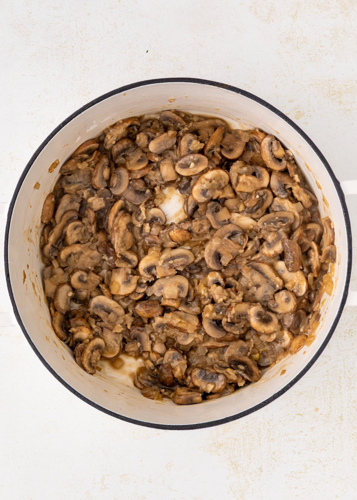 Sliced mushrooms cooking in a white pot on a light-colored surface.