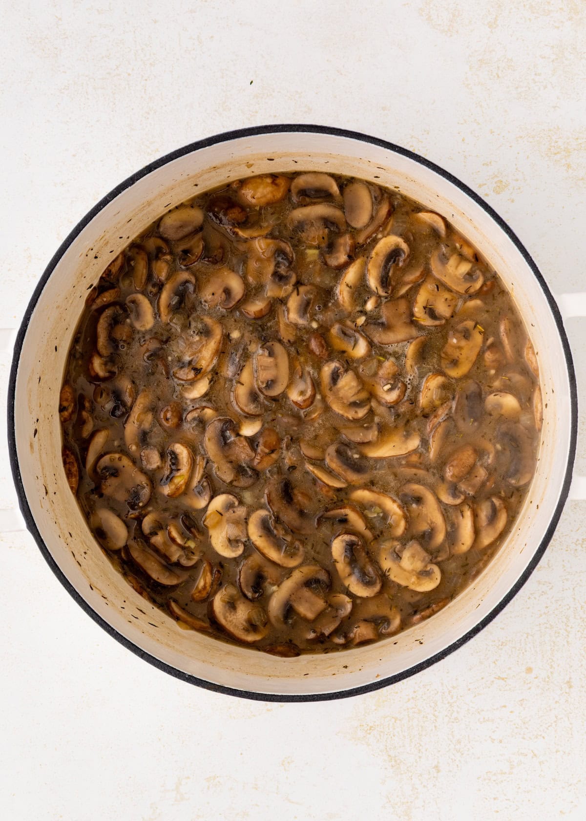 Sliced mushrooms cooking in a large white pot with broth on a light surface.