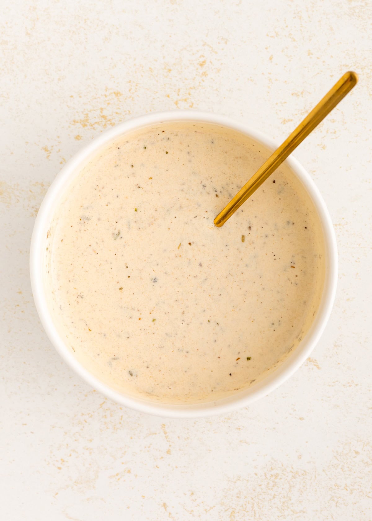 Creamy Italian dressing speckled sauce in a white bowl with a gold spoon on a light background.