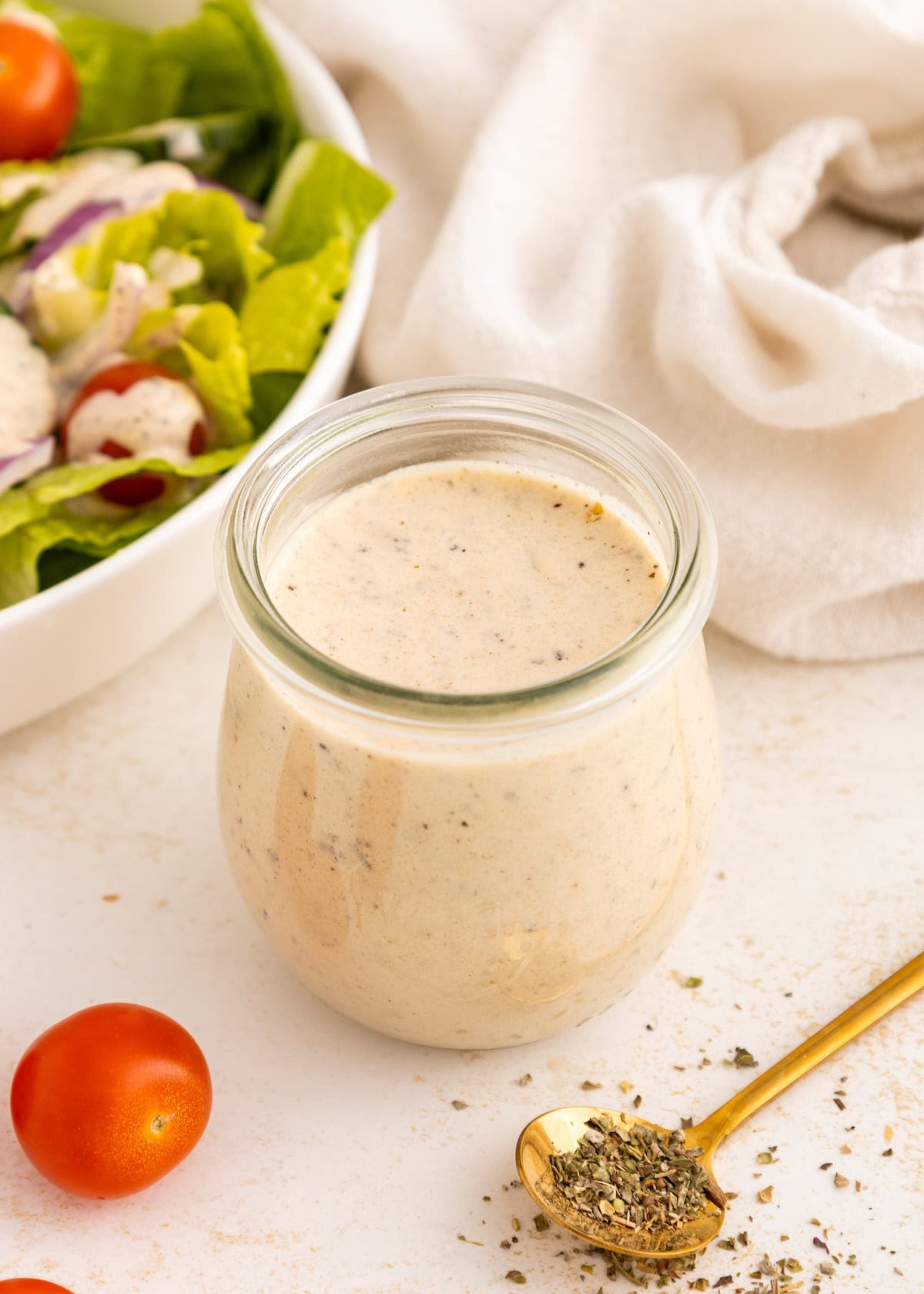 Glass jar of creamy Italian dressing beside a salad, cherry tomatoes, and a gold herb spoon.