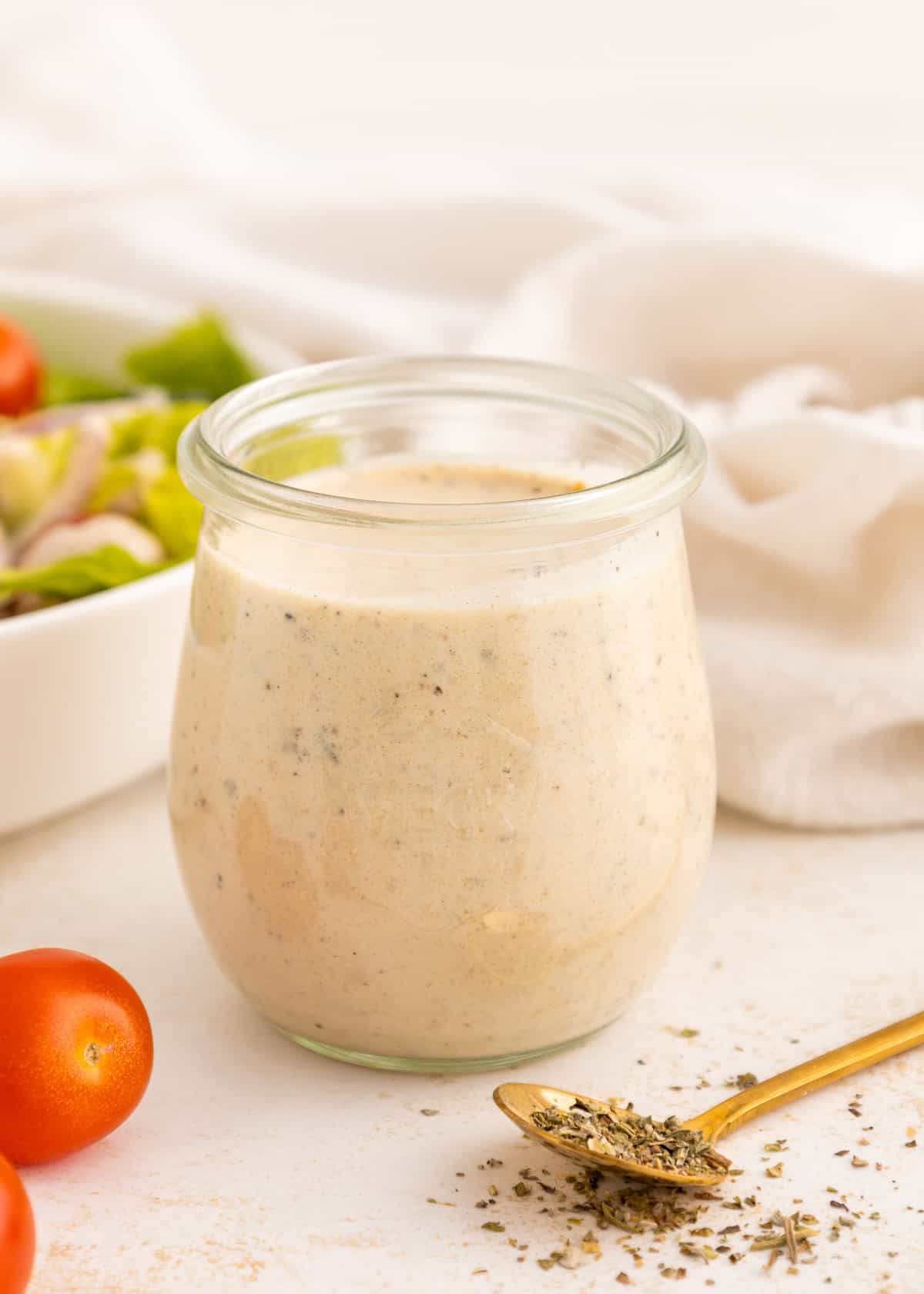 A glass jar of creamy Italian dressing with herbs, next to a gold spoon and cherry tomatoes.