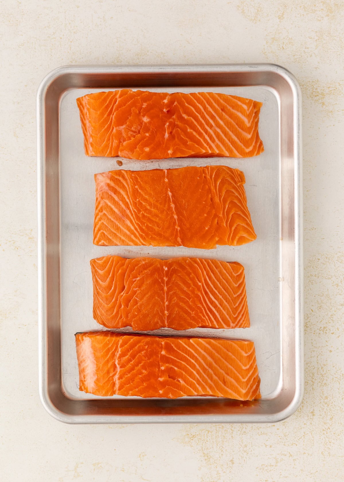 Four raw salmon fillets arranged in a row on a metal baking tray.
