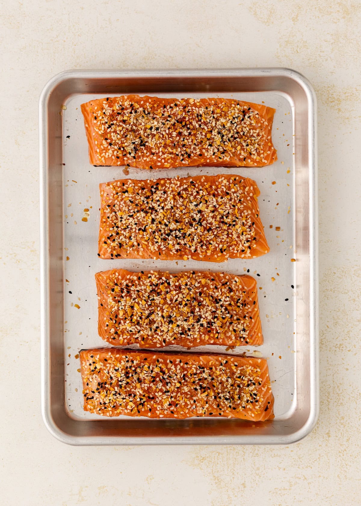 Four seasoned salmon fillets on a metal baking tray, viewed from above.
