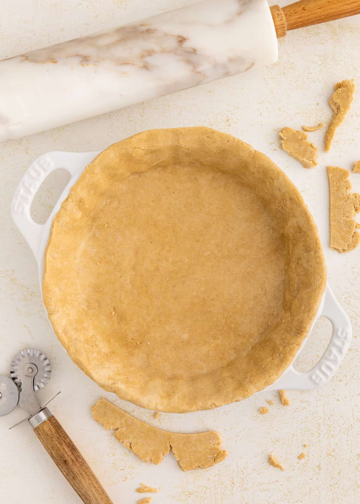 Unbaked pie crust in a white dish with a marble rolling pin and pastry cutter nearby.